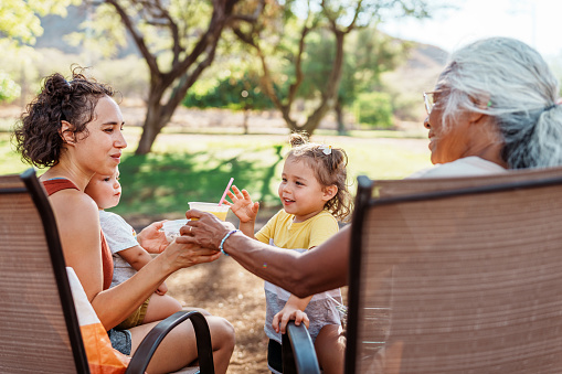 A senior woman of Pacific Islander ethnicity shares a smoothie with her adult daughter during a picnic outside on a beautiful day in Hawaii.