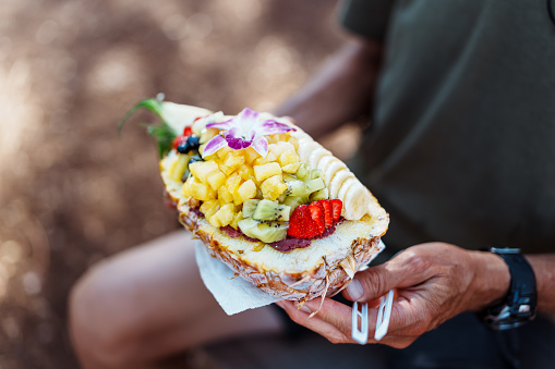 An unrecognizable man holds a pineapple açaí bowl filled with fresh fruit during an outdoor picnic on a beautiful weekend in Hawaii.