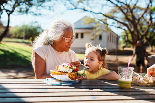A senior woman of Pacific Islander ethnicity sits at a picnic table with her multiracial toddler granddaughter, helping her eat an açaí bowl outside.