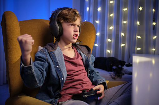 Expressive curly haired boy in headphones sitting on comfortable armchair with gamepad while playing videogame and celebrating victory with raised arm and clenched fist near sleeping adorable Miniature Schnauzer