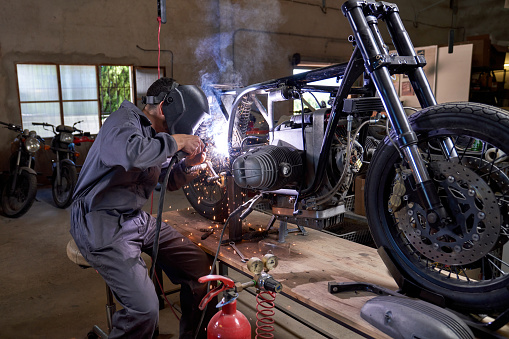 Side view of anonymous repairman in protective helmet using welding machine while fixing disassembled motorcycle in workshop
