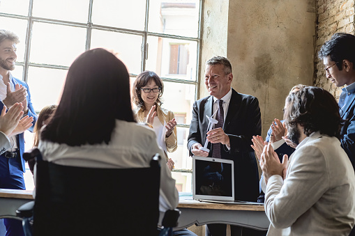 A diverse group of people, standing and sitting around a table applauding a man holding a model of a wind turbine, in an office. Celebrating teamwork and innovation in the renewable energy industry.