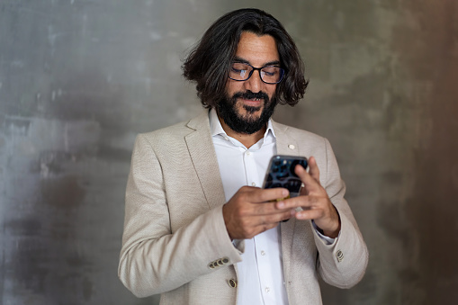 A young Arab businessman in shirt and beige jacket with long hair and beard, smiles using a smartphone in a neutral office setting with an antiqued wall - the phone screen reflects on his glasses