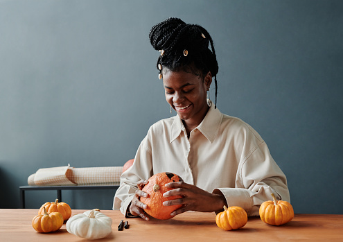 Young smiling African American woman looking at spooky face on ripe Halloween pumpkin drawn with black highlighter
