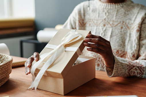 Hand of young unrecognizable black woman in warm sweater opening giftbox with tied white ribbon on top of cover while sitting by table