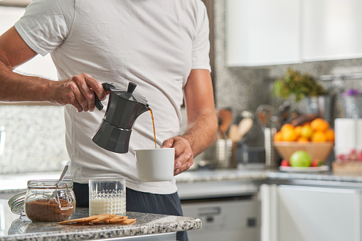 Crop anonymous male in casual clothes pouring hot coffee from geyser coffeemaker into mug while preparing breakfast in light kitchen