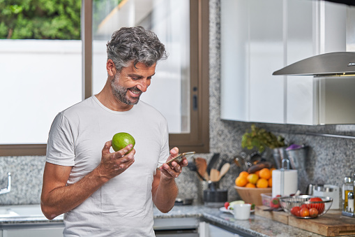 Cheerful middle aged bearded man with gray hair standing in kitchen with green apple and texting message on cellphone