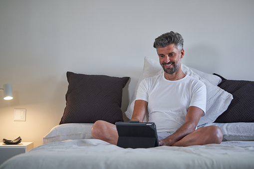 Mature male entrepreneur in sleepwear smiling and looking at screen of tablet while working on project remotely in bedroom at home