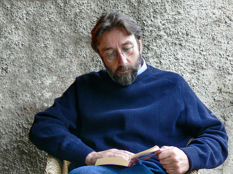 A bearded man sits on a chair in front of a gray wall and reads a book intently
