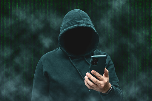 Mysterious faceless hooded anonymous hacker holding smartphone, silhouette of cybercriminal, terrorist.
