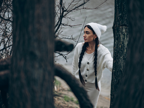 Young dreamy woman with braided hair standing in forest near trunk tree in white sweater, skirt and hat.