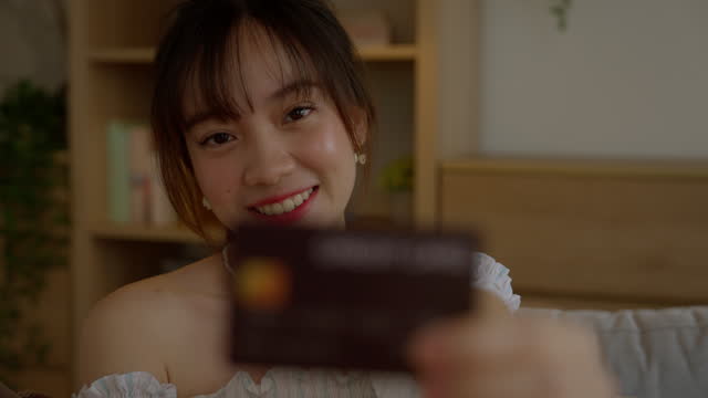 Happy smiling young Asian woman showing credit card and looking camera at home.