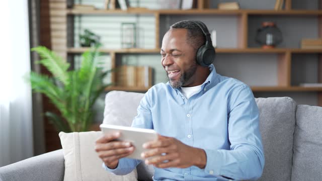 Excited mature african american male in wireless headphones watching sports match competition on tablet sitting on sofa in living room at home