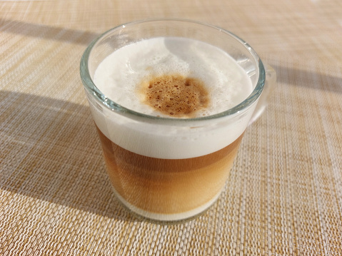 Close-up shot of hot cappuccino coffee in a glass mug with top layer of frothy steamed milk foam and espresso made from coffee capsules on a table