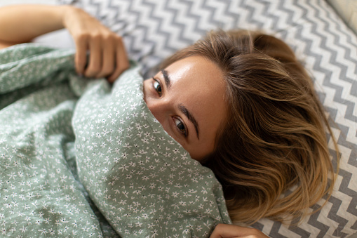 A beautiful young woman lying in her bed, covering her face with duvet, looking happy