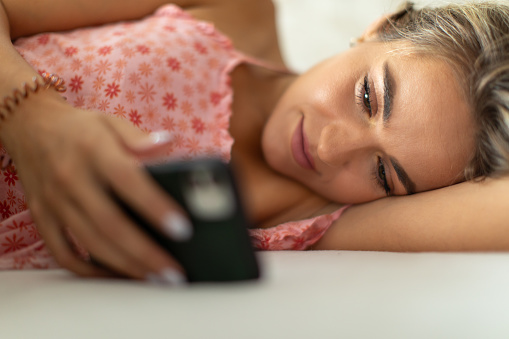A beautiful young woman lying on side in her bed, looking and typing on her phone, she looks happy, with a smile on her face