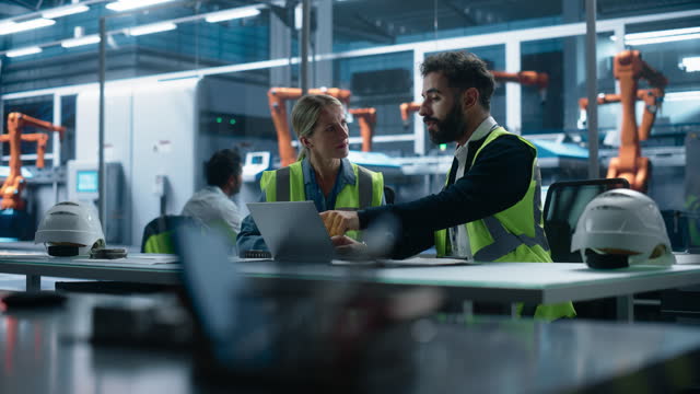 Autonomous Factory Office: Caucasian Female Quality Control Technician Talks to Hispanic Male Industrial Engineer Working on Laptop Computer. Automated Robot Arm Assembly Line Manufacturing Machinery.