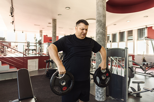 A middle aged man is exercising in a gym, he is lifting barbell plates, his aim is to lose weight