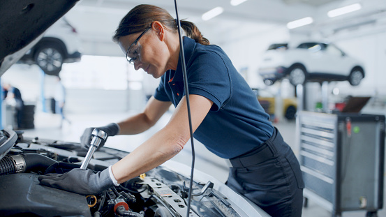 Expert Female Mechanic Turning A Socket Wrench Under The Car Engine Hood At Auto Repair Shop