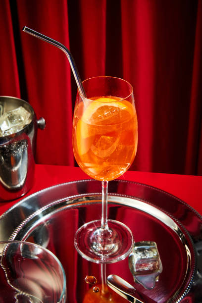 foto ravvicinata di un bicchiere alto riempito di cocktail all'arancia, aperol con pezzo di agrumi e paglia seduto su un vassoio d'argento con cubetto di ghiaccio. - martini brand vermouth foto e immagini stock