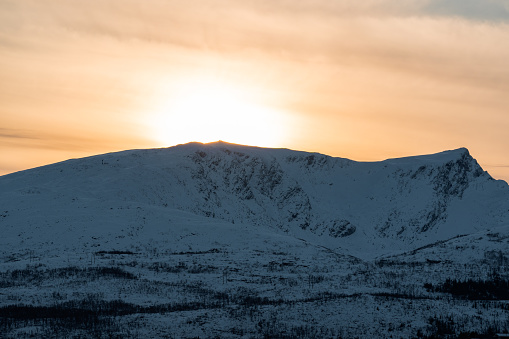 Golden suunseet behind a Norweegian mountain during winter, sun peering through the cloudcover to leave a golden haze