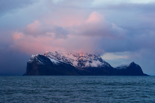 Purple clouds above a mysterious island covered in snow in the ocean, hues of red and blue clouds over the turquoise ocean, clouds covering the peak of the mountains
