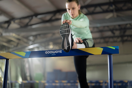 Athlete young woman stretching on hurdle in sports hall.