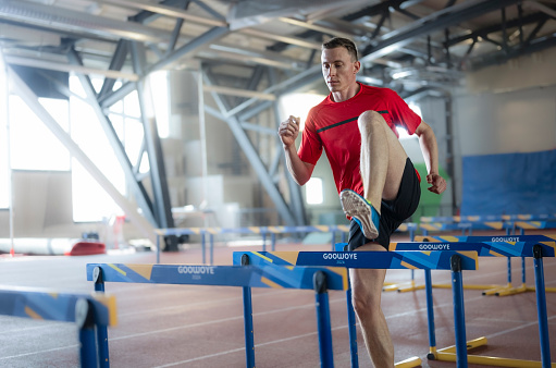 Athlete man doing warm-up exercises on hurdle track in sports hall.