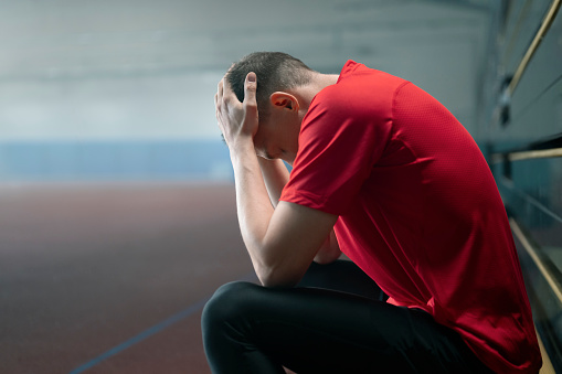 Side view of stressed gymnast man sitting with head in hands in sports hall.