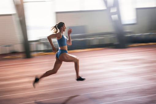 Blurred motion of young female sprinter running on indoor track in sports hall during practice.