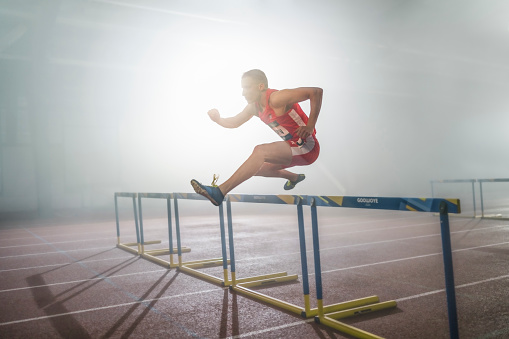 Athlete man jumping over hurdles during practice in sports hall.