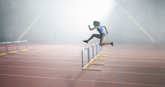 Side view of athlete young woman jumping over hurdles during practice in sports hall.