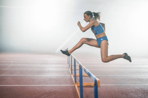 A female collegiate heptathlete jumps hurdles during a one-on-one training session with her coach.