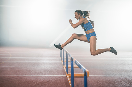 Side view of athlete young woman jumping over hurdles during practice in sports hall.