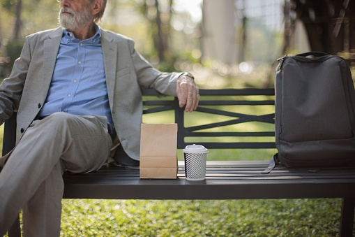 Midsection of a senior businessman sits on the bench in park with a takeaway coffee and breakfast by his side.