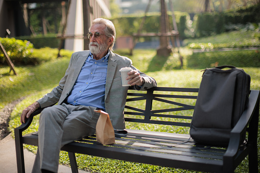 Senior businessman sits pensively on the bench in park holding a paper coffee cup in hand.