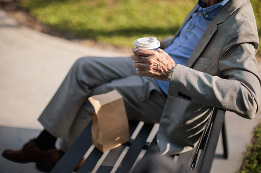 Midsection of a senior businessman sits on the bench holding a paper coffee cup during a breakfast break in the park.