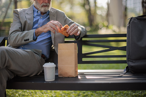 Midsection of a senior businessman sits on the bench in park, enjoying breakfast.