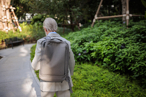 Rear view of a senior businessman with a backpack walking through the park.
