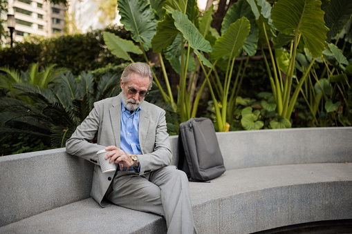 A senior businessman is checking the time on a hand watch while relaxing on a concrete bench during a work break.