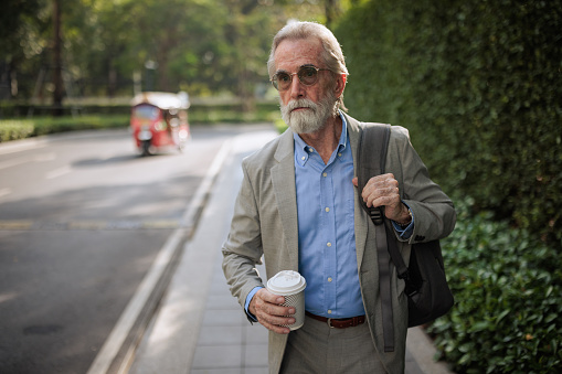 A senior businessman walks along the sidewalk near the street, holding a takeaway coffee.