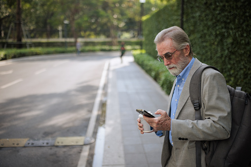 A senior businessman checks the taxi location on the app while waiting on the street.