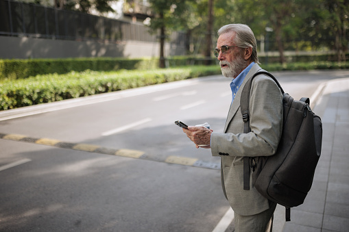 A senior businessman checks the taxi location on the app while waiting on the street.