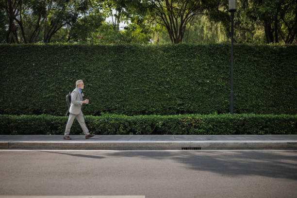 Senior businessman walking with a backpack A senior businessman walks along the sidewalk near the street, holding a takeaway coffee. bangkok street stock pictures, royalty-free photos & images