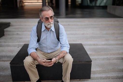 A senior businessman is sitting pensively with a phone in his hand on a concrete bench in the city.