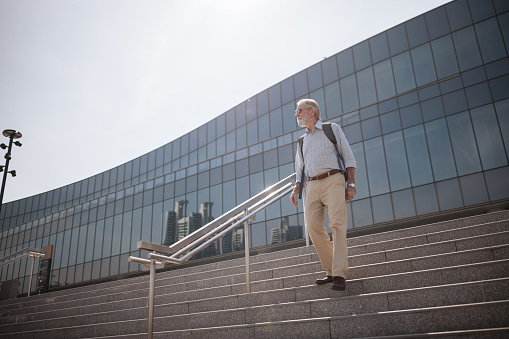 A senior businessman with a backpack walks down the stairs in front of the office building.