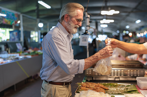A smiling senior man is buying street food at the food court in Bangkok.