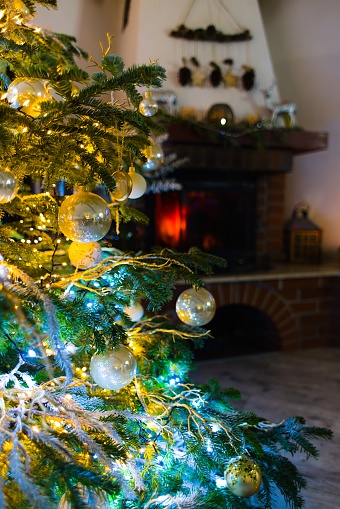 Immerse in the holiday spirit with this enchanting photo featuring baubles on a Caucasian Fir. Blurred Christmas lights and festive decor enhance the cozy atmosphere, while a red brick fireplace adds warmth and charm to the scene.