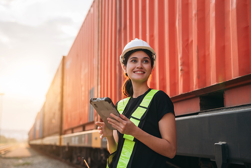 Foreman checking inventory or task details on freight train cars and shipping containers. Logistics concept, import, and export industries.