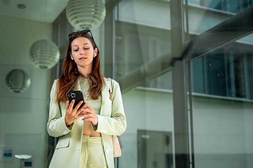 Beautiful young woman stands against office buildings wearing an elegant green suit while checking her phone